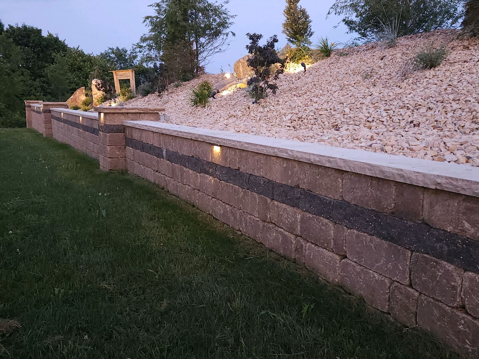 Stone retaining wall along a grassy slope with white rock landscaping and small lit trees at dusk
