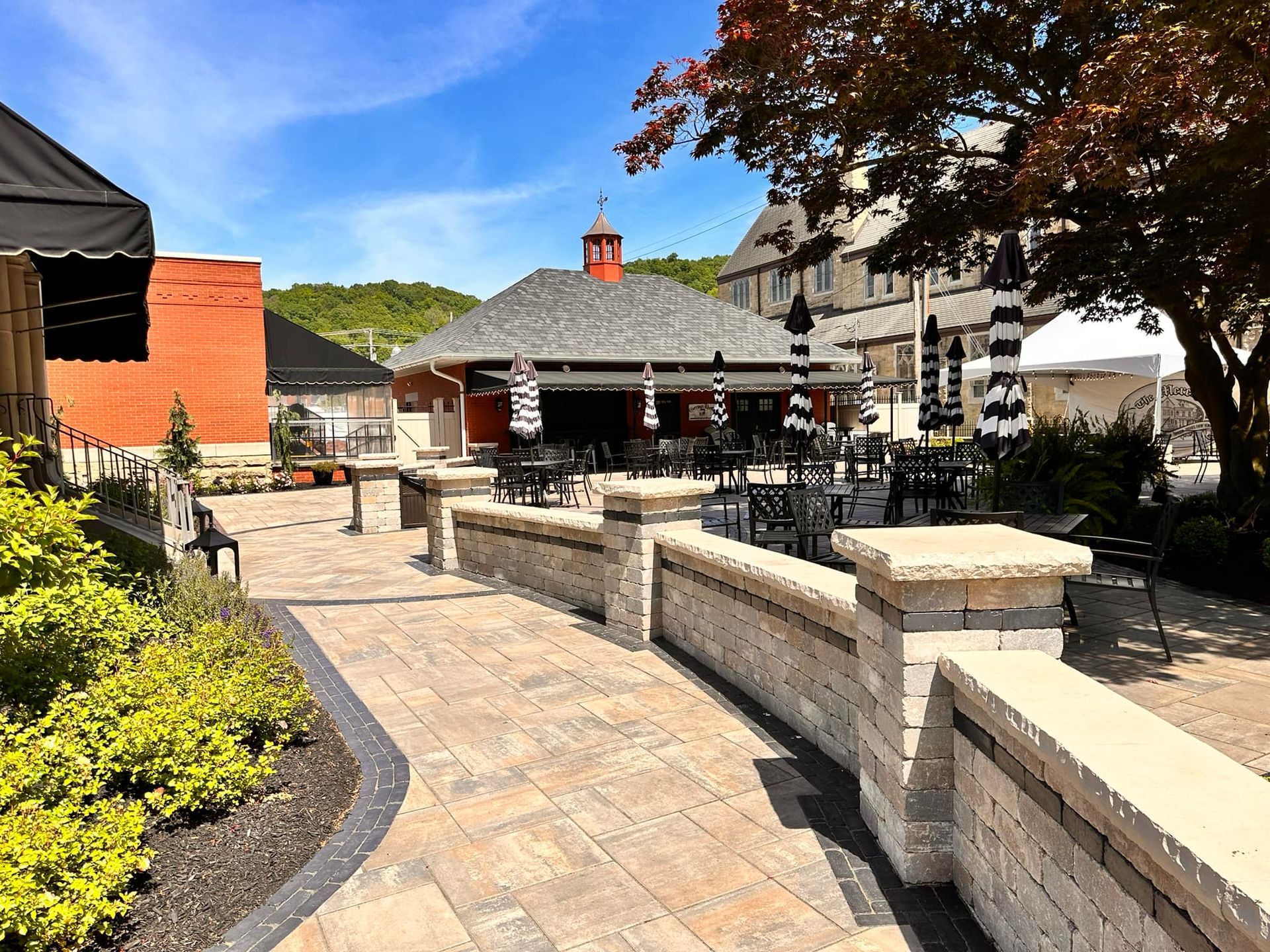 Outdoor patio with stone seating, tables, and umbrellas beside buildings under a bright blue sky