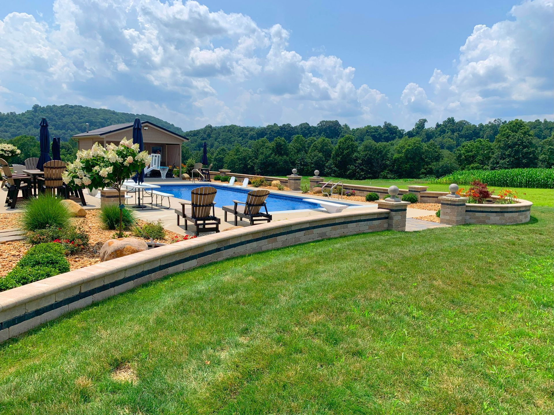 Backyard pool with lounge chairs, stone patio, and green lawn under a partly cloudy sky