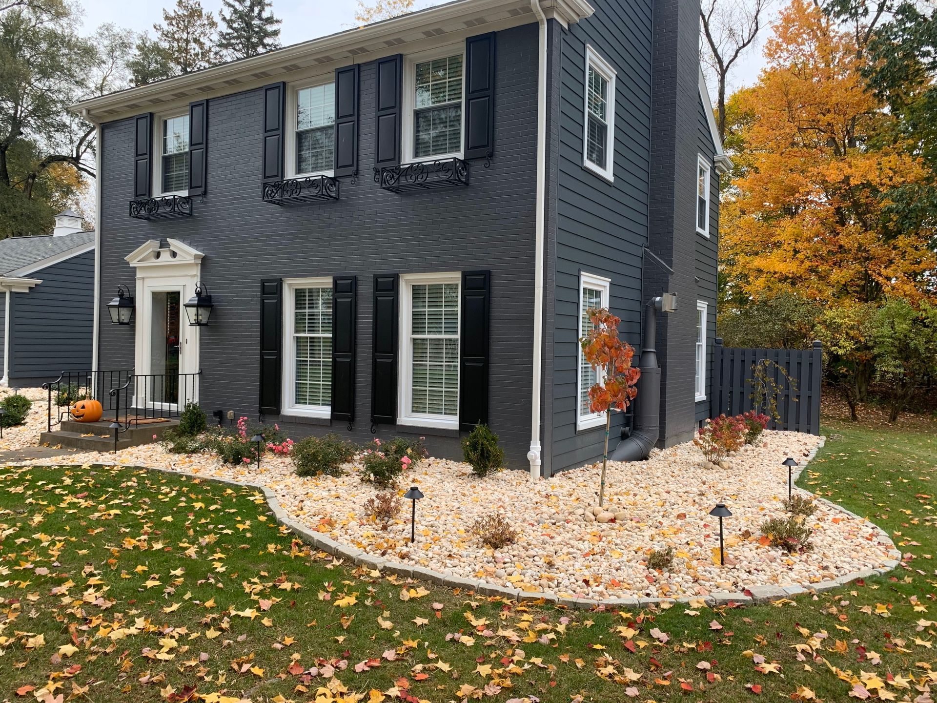 Two-story dark gray house with black shutters and white trim, bordered by a white stone garden bed in autumn.