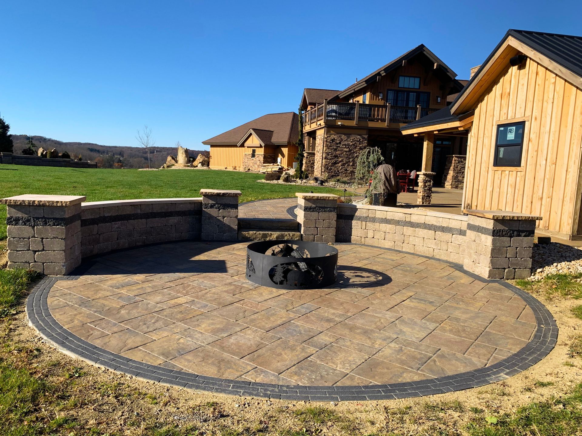 Stone patio with circular fire pit, built-in benches, and a rustic wooden house in the background.