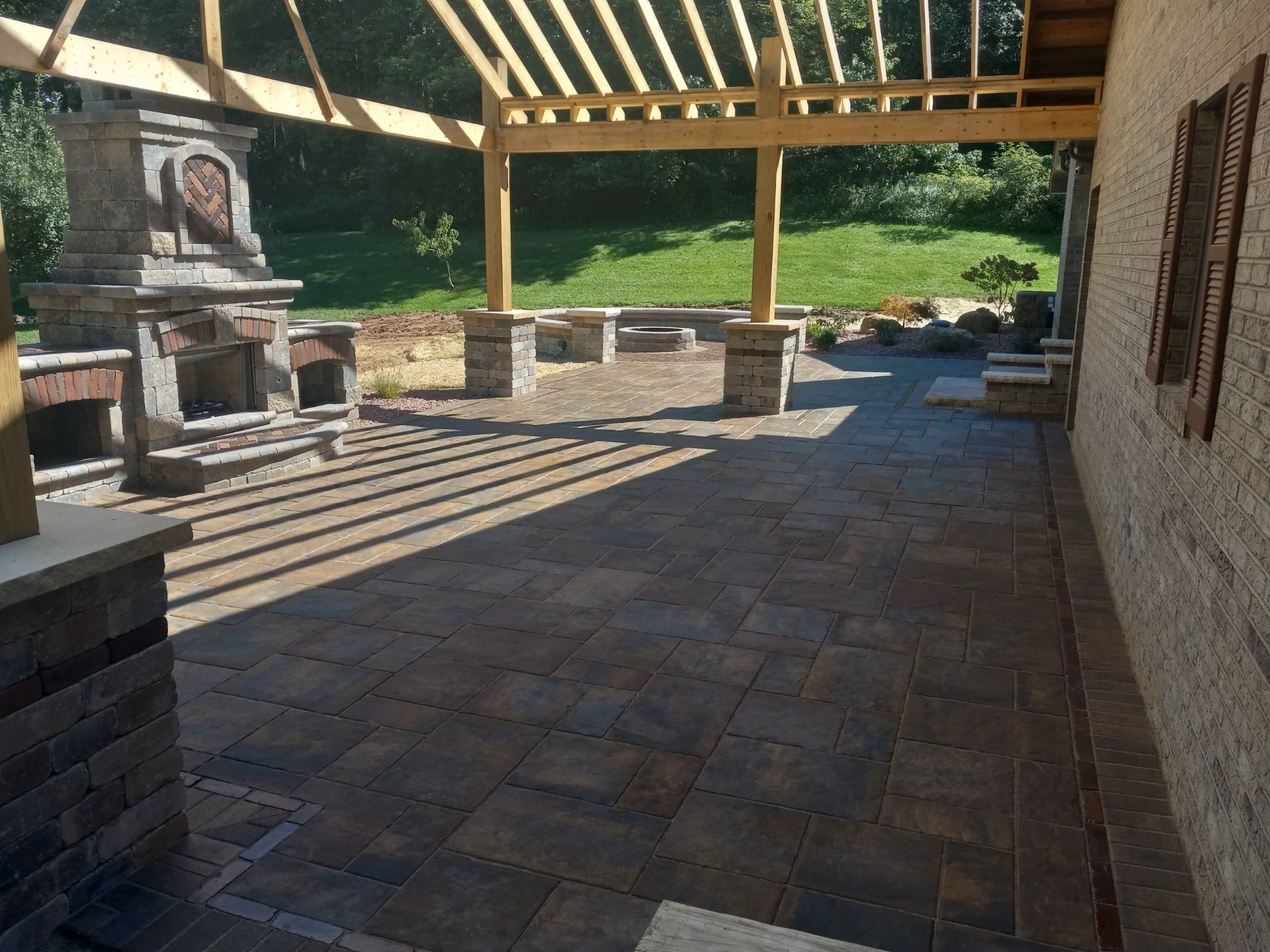 Covered stone patio with wooden pergola, brick fireplace, and empty outdoor seating area in sunlight