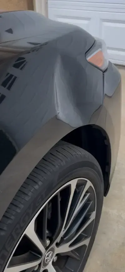 Black car with a dented rear fender near a garage door.