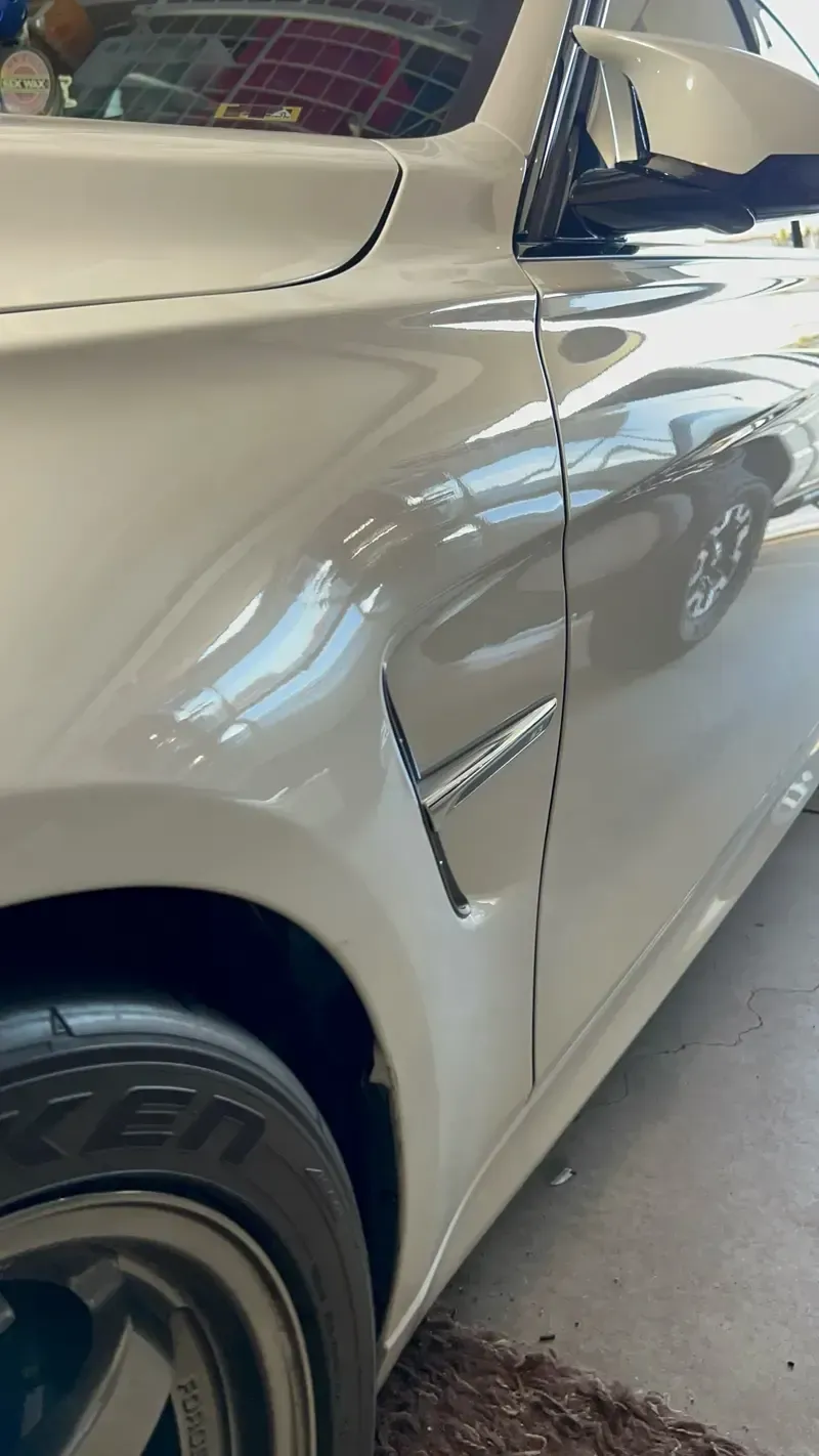White car's front quarter panel with a side vent, a black tire, and a partial view of its side mirror.