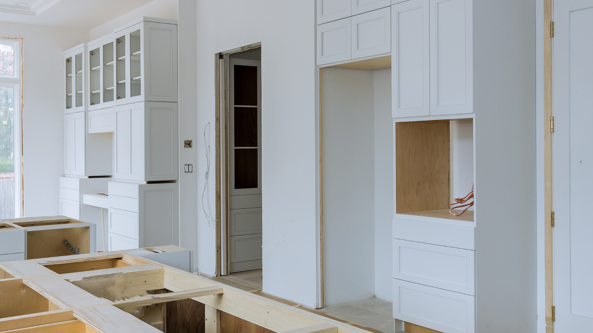 Bright unfinished kitchen with white cabinets, open doorways, and exposed floor framing in a renovation.
