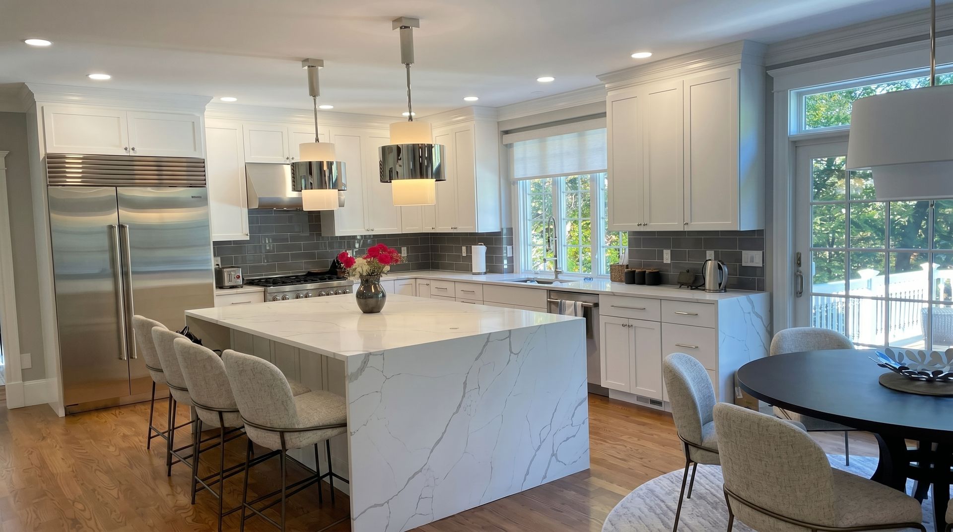 Bright modern kitchen with white island, stainless fridge, pendant lights, and round dining table by French doors