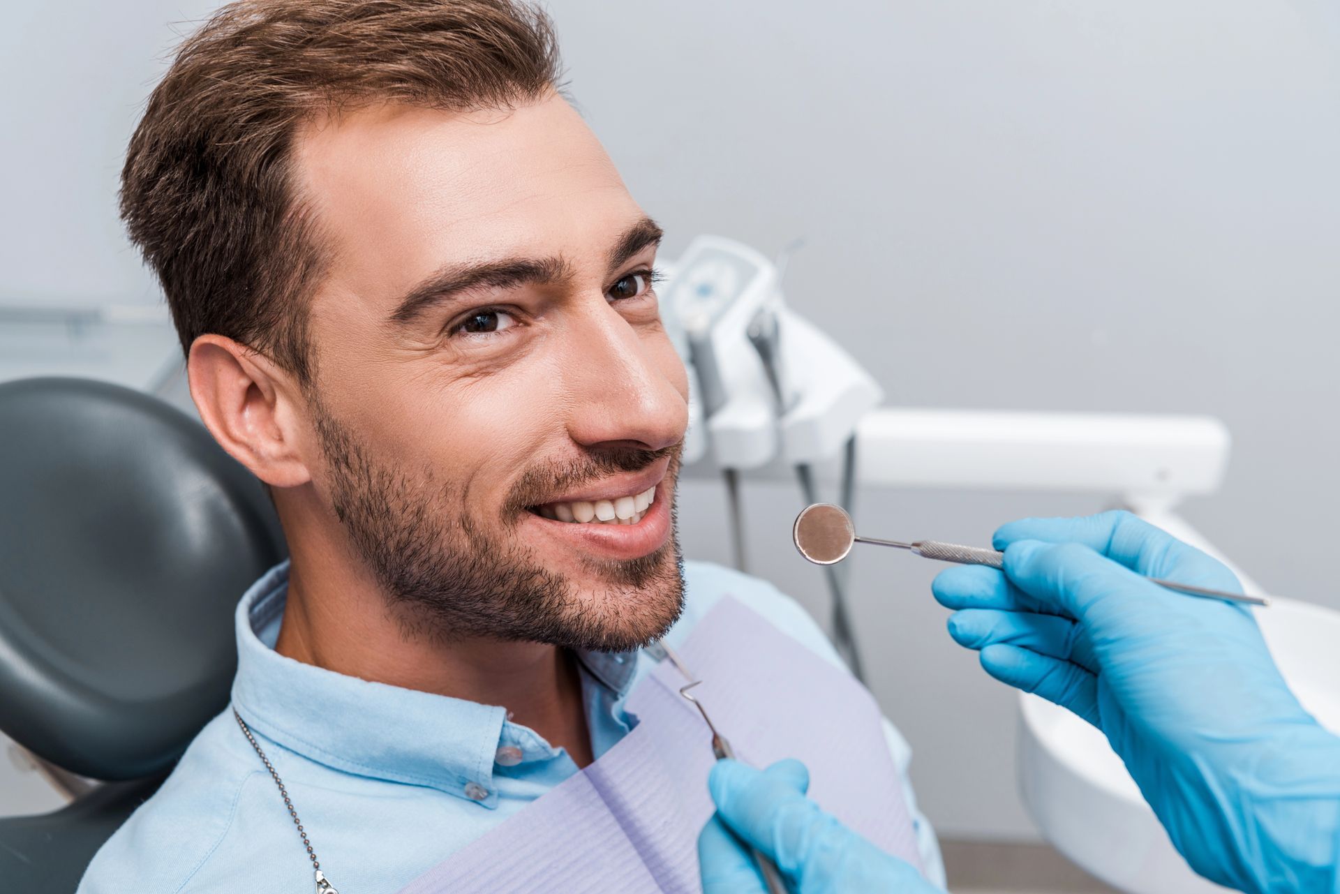 Una persona sorride seduta sulla poltrona del dentista, mentre un professionista con i guanti blu tiene uno specchietto dentale.