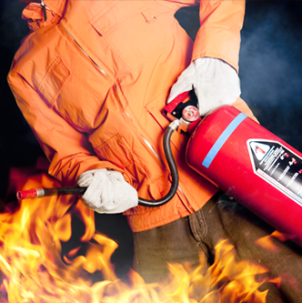 Persona con giacca arancione che usa un estintore per spegnere un incendio, indossando guanti.