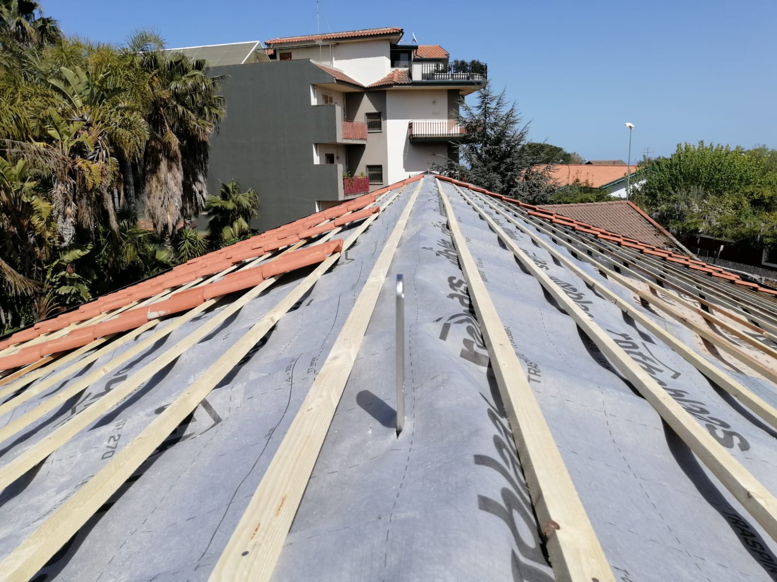 Tetto in costruzione con travi in legno, membrana grigia e tegole arancioni contro il cielo azzurro.