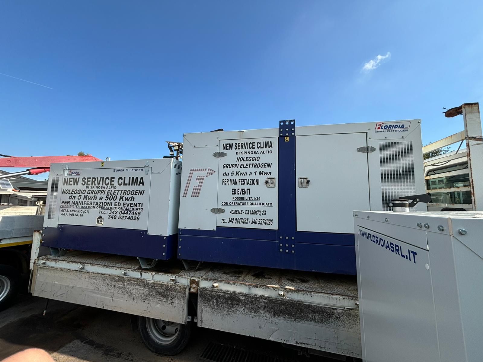 Generatori sul cassone di un camion, blu e bianchi con scritte nere. Cielo limpido sullo sfondo.