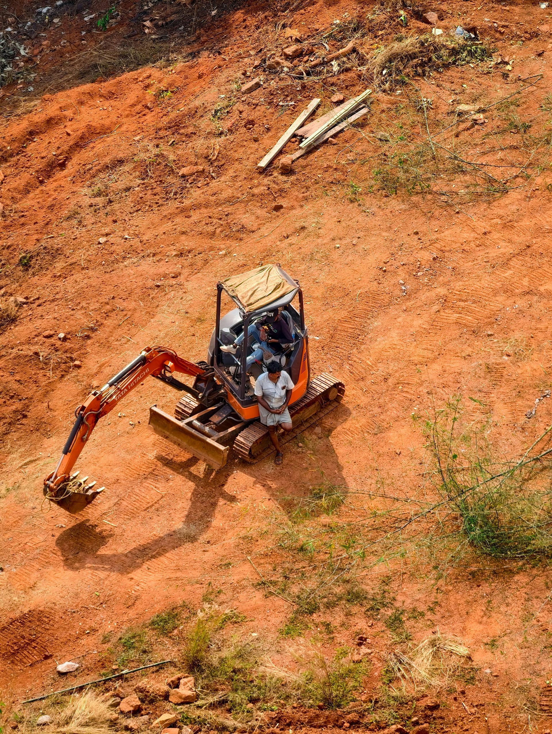 Un piccolo escavatore arancione su una collina di terra rossastra. Due persone sono sedute all'interno.