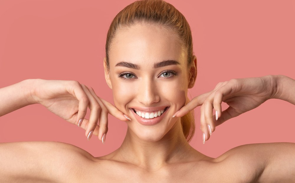 Woman smiling, fingers pulling cheeks, fair skin, pink background.