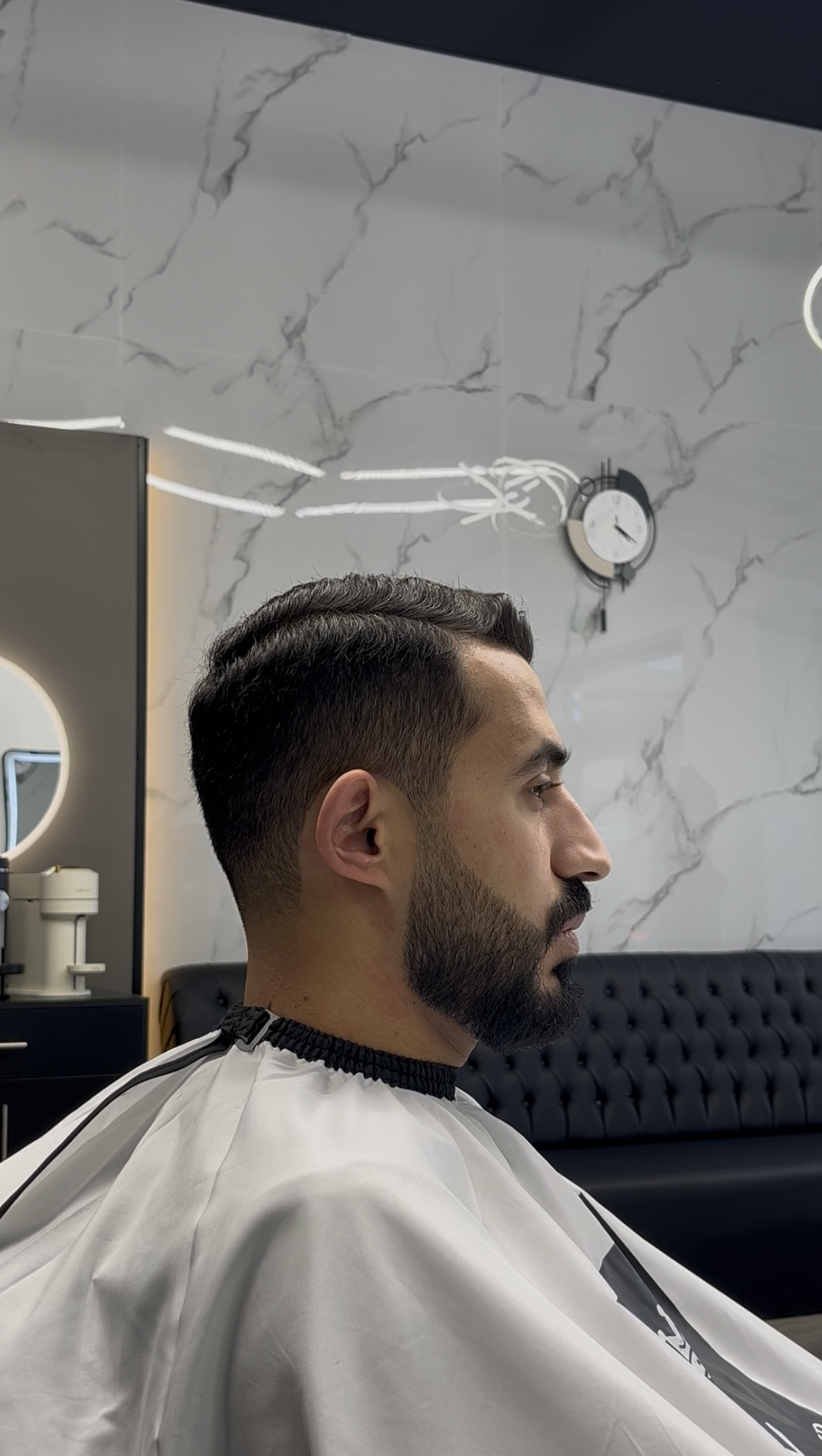 Man getting a haircut in a barber shop, side profile view. He is wearing a barber cape.