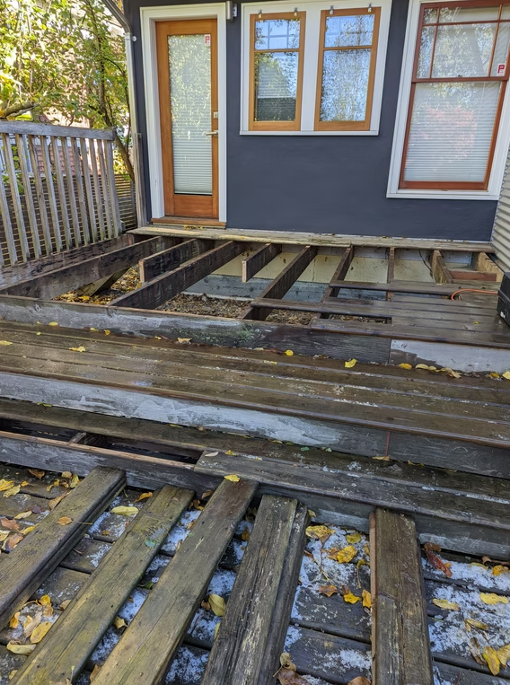 Damaged wooden deck with missing boards beside a gray house and glass door