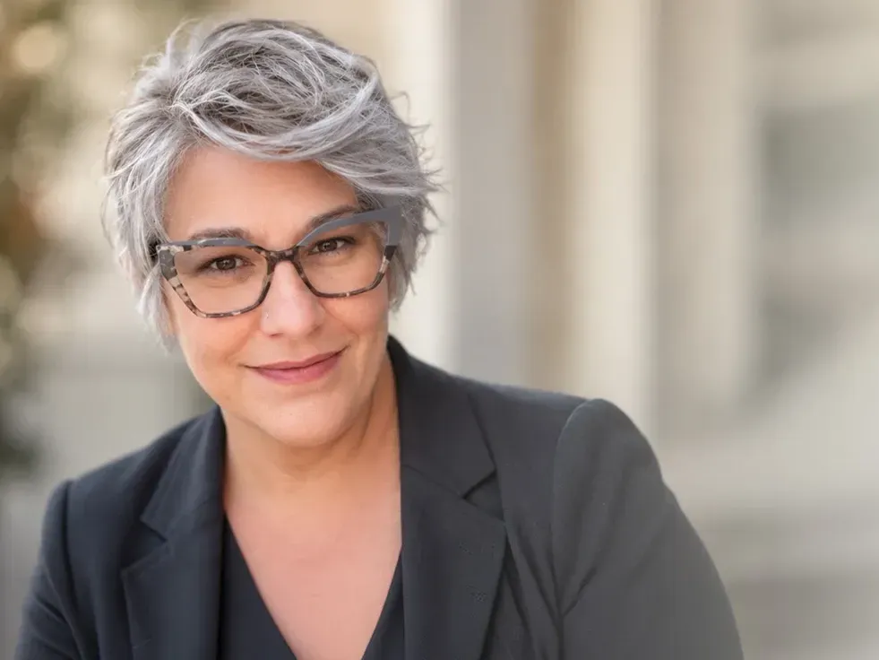 Smiling person with short gray hair and glasses in a black blazer against a warm, blurred background
