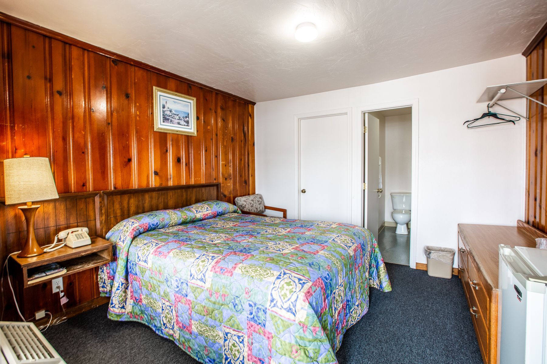 A hotel room with a bed , dresser , lamp , and toilet.