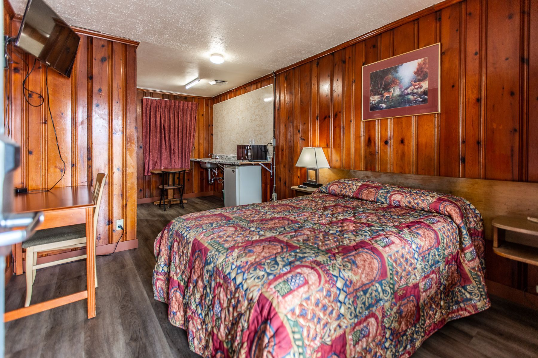 A hotel room with a bed , desk , television and wood paneling.