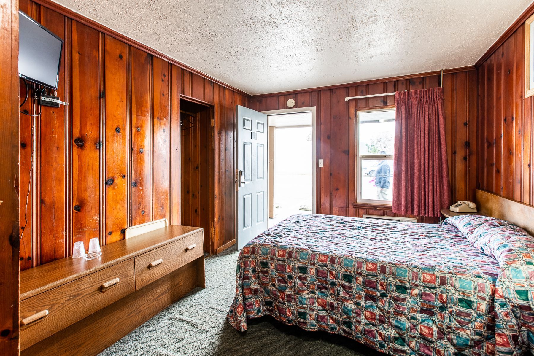 A hotel room with a bed , dresser , television and wood paneling.