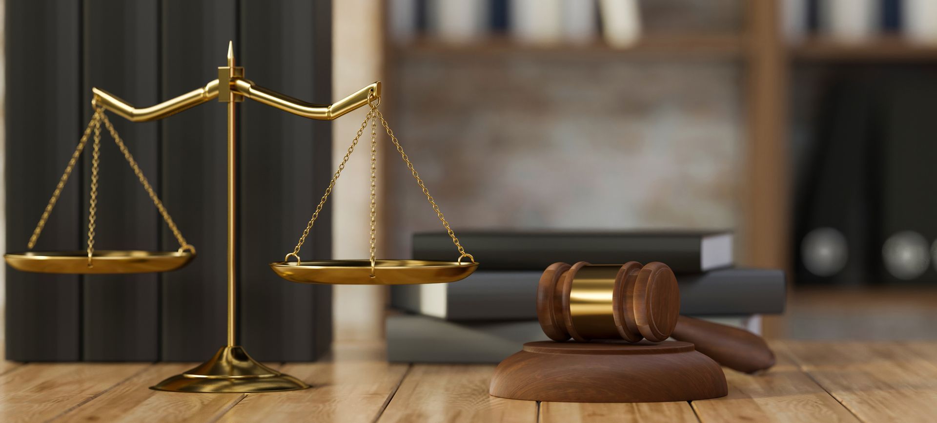A gold balance scale and a wooden gavel resting on a desk in front of a blurred bookshelf filled with law books.