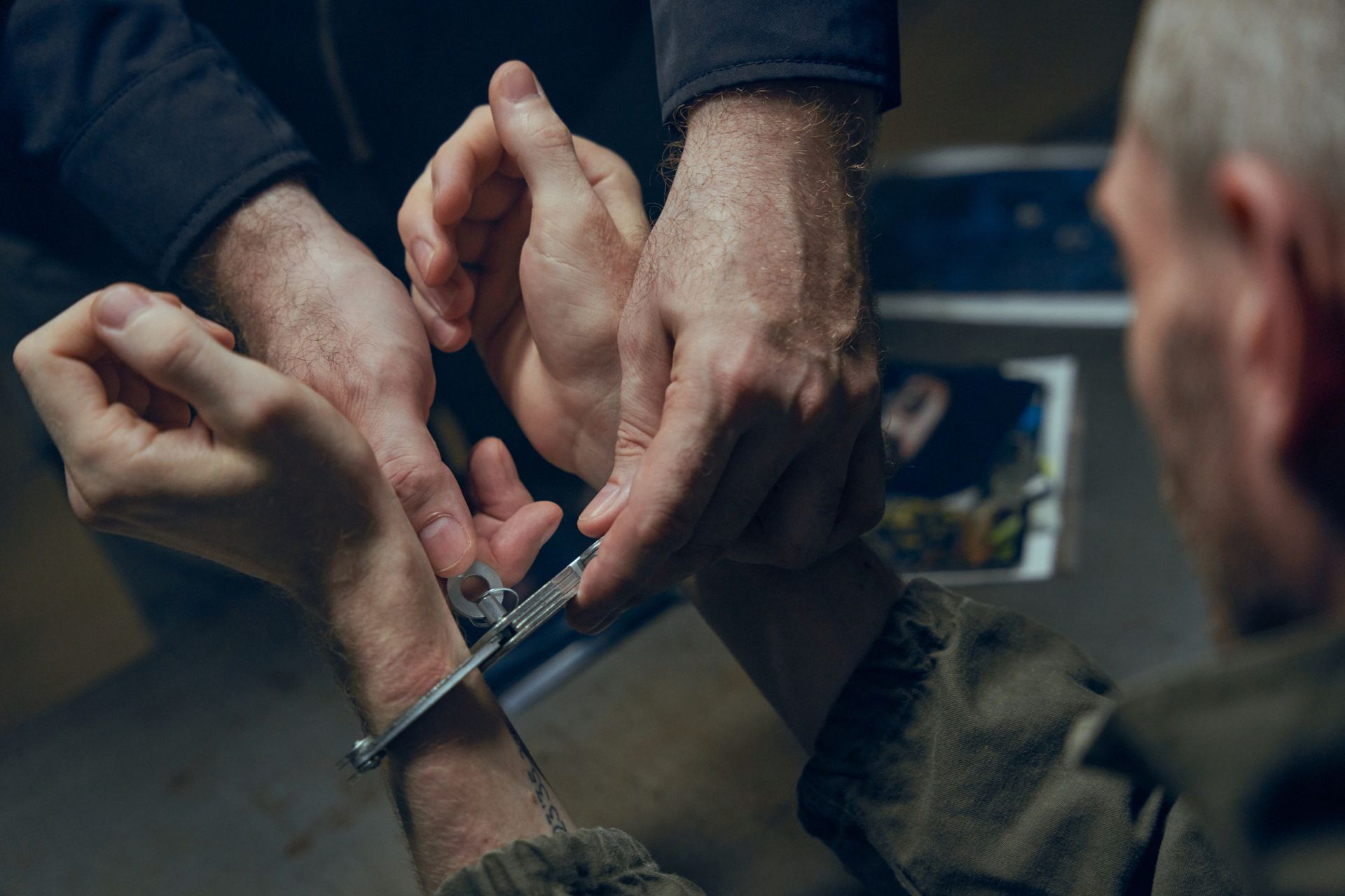 Hands with handcuffs held by an officer over a desk with documents in a dimly lit room.