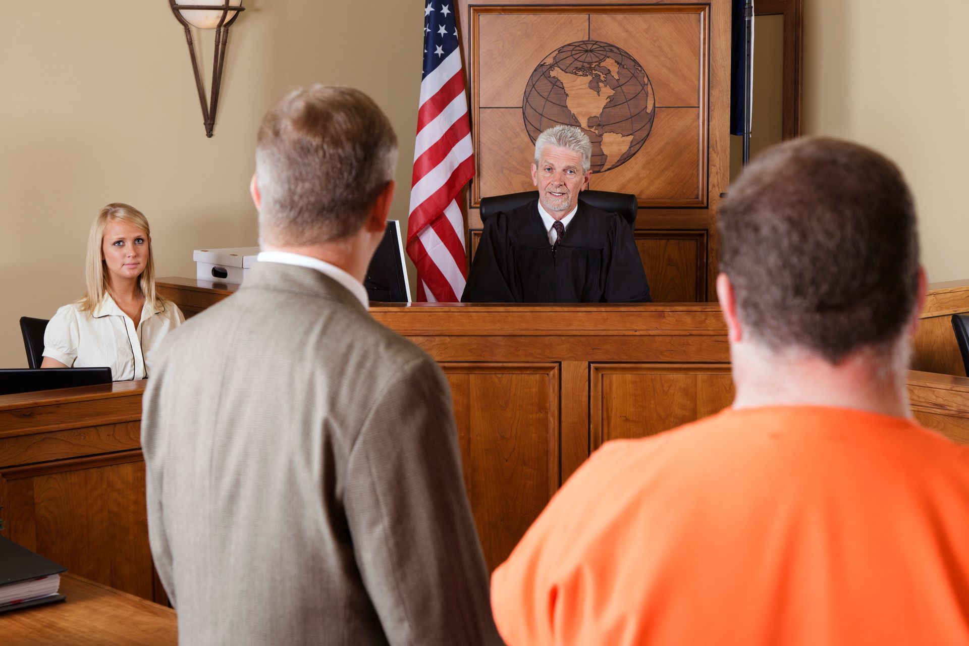 Male lawyer stands next to man in jail clothes in front of a judge and a witness, inside a courtroom.