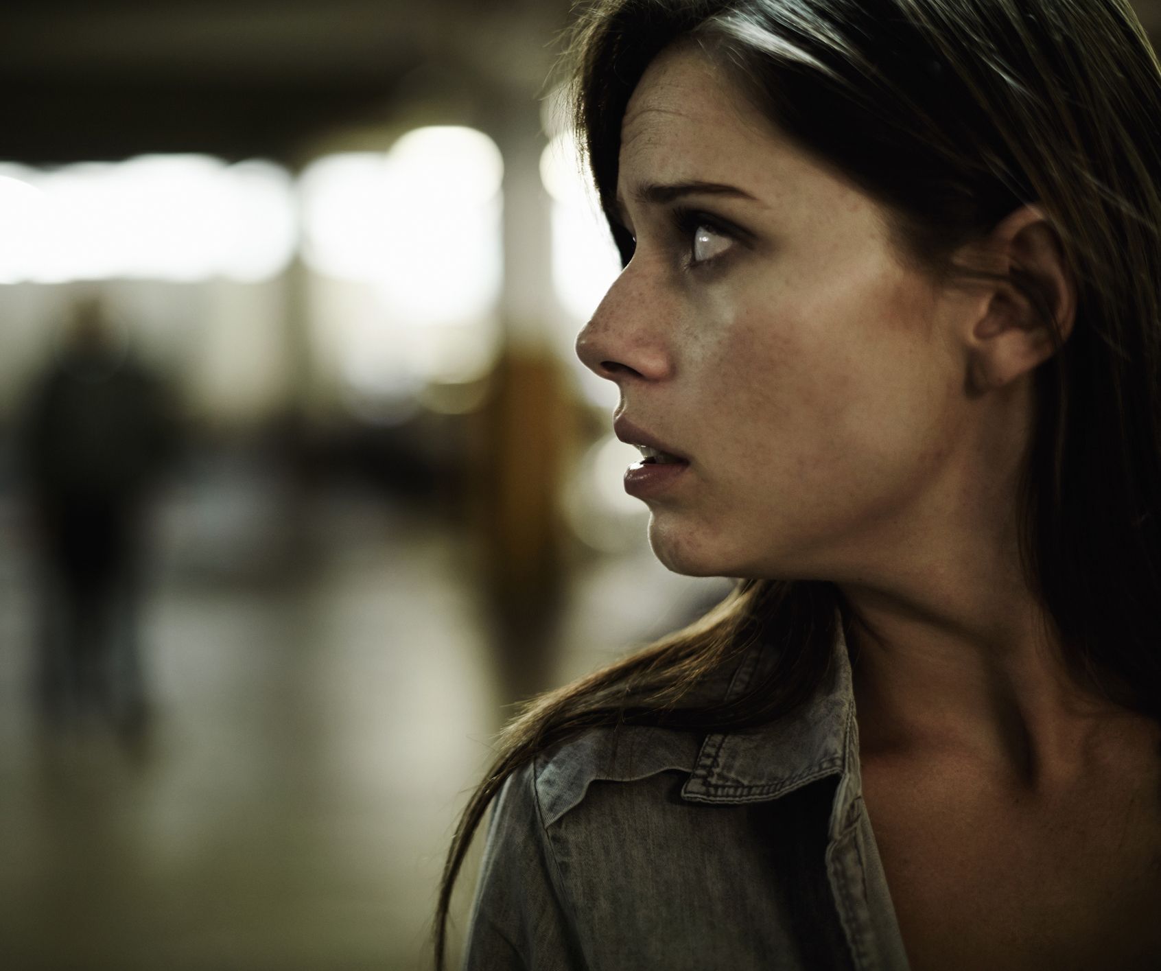 A person with a wide-eyed, anxious expression looks over their shoulder in a dimly lit, out-of-focus indoor space.