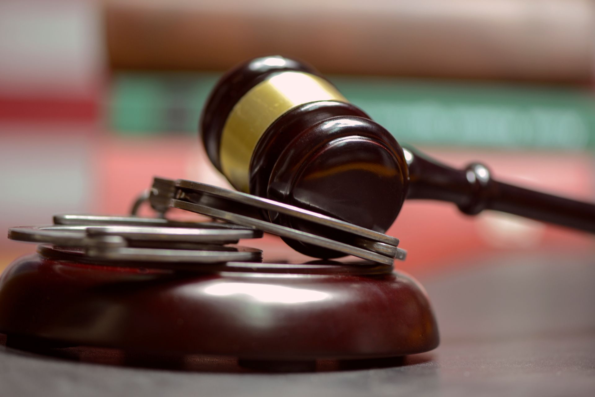 A wooden gavel rests on its block next to a pair of silver handcuffs, with out-of-focus books in the background.