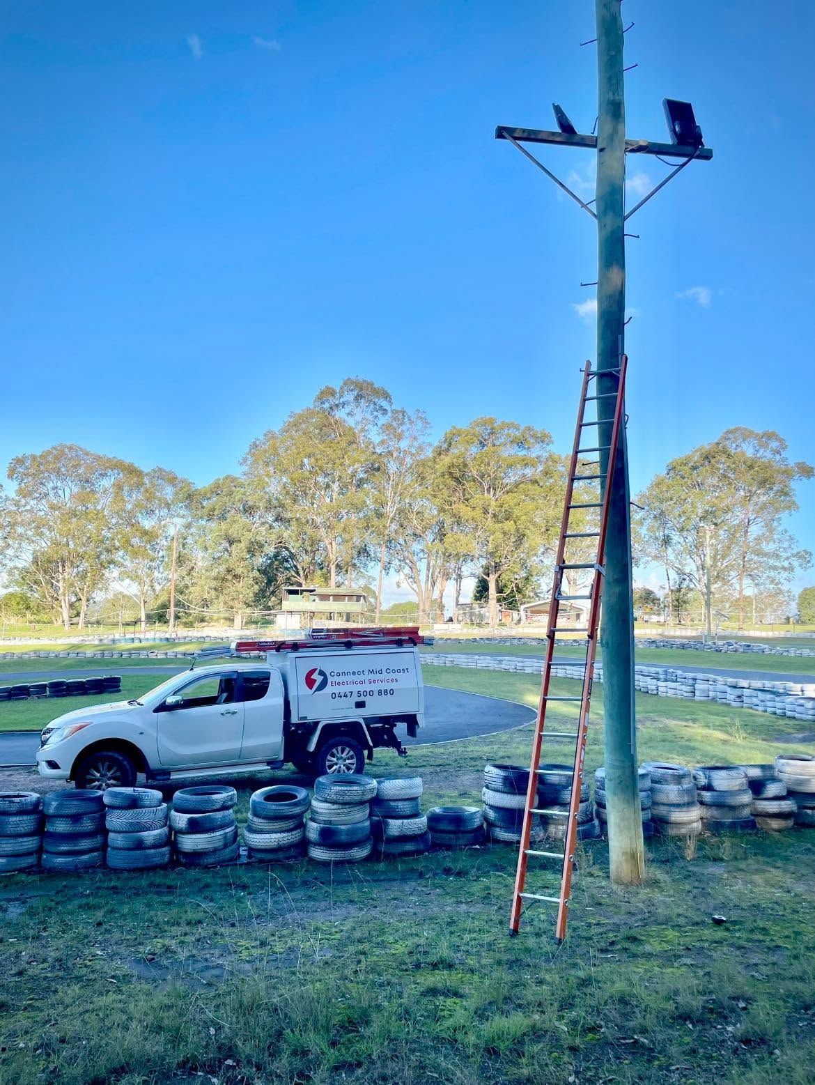 White utility truck beside stacked tires; ladder leans on a utility pole with a worker at the top. — Connect Mid Coast Electrical Services In Taree, NSW