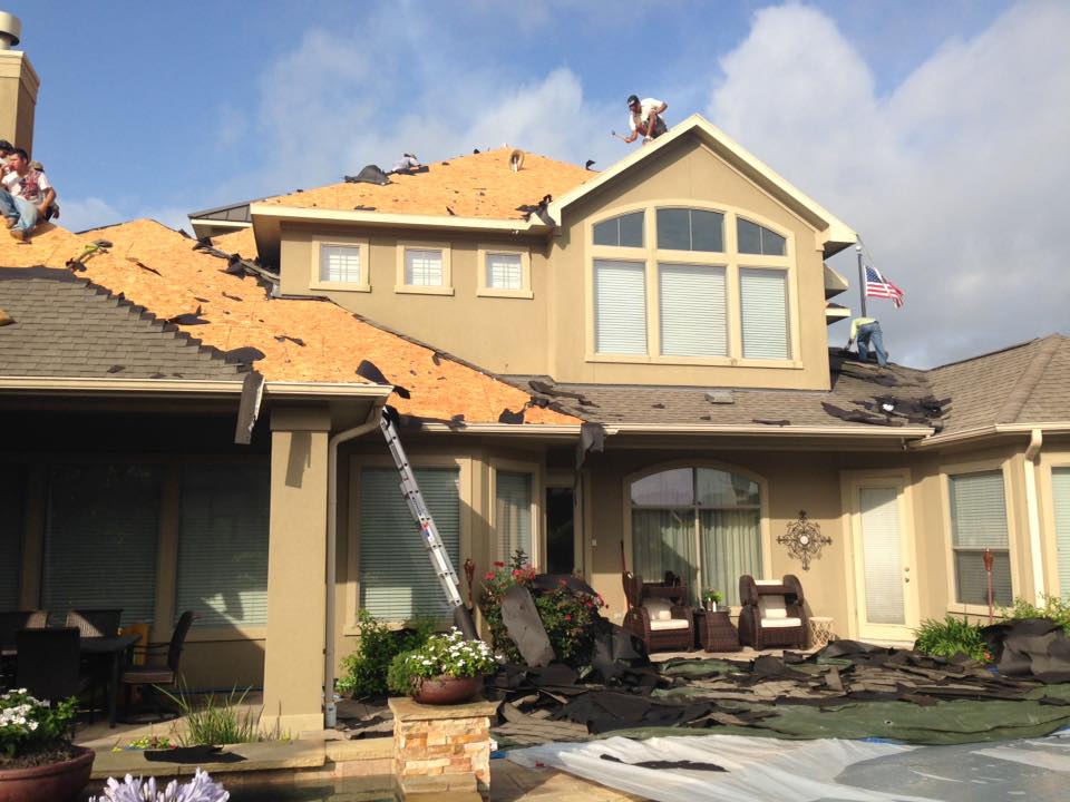 Workers strip old shingles off a large, multi-story house roof, exposing the wooden deck.