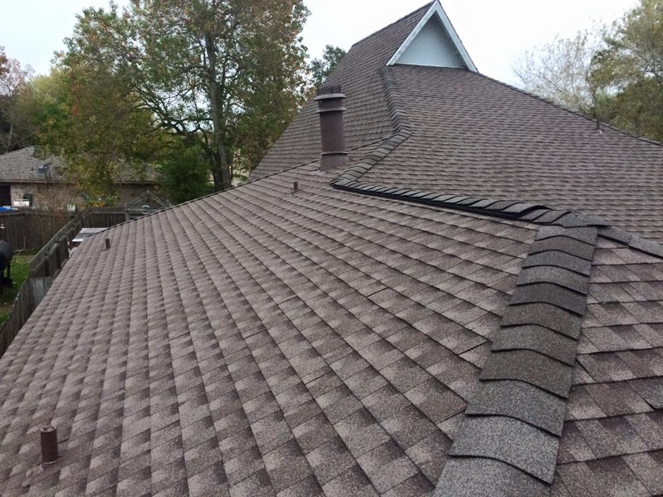 A high-angle view of a brown shingled roof with a chimney, a roof vent, and a peak against a backdrop of trees.