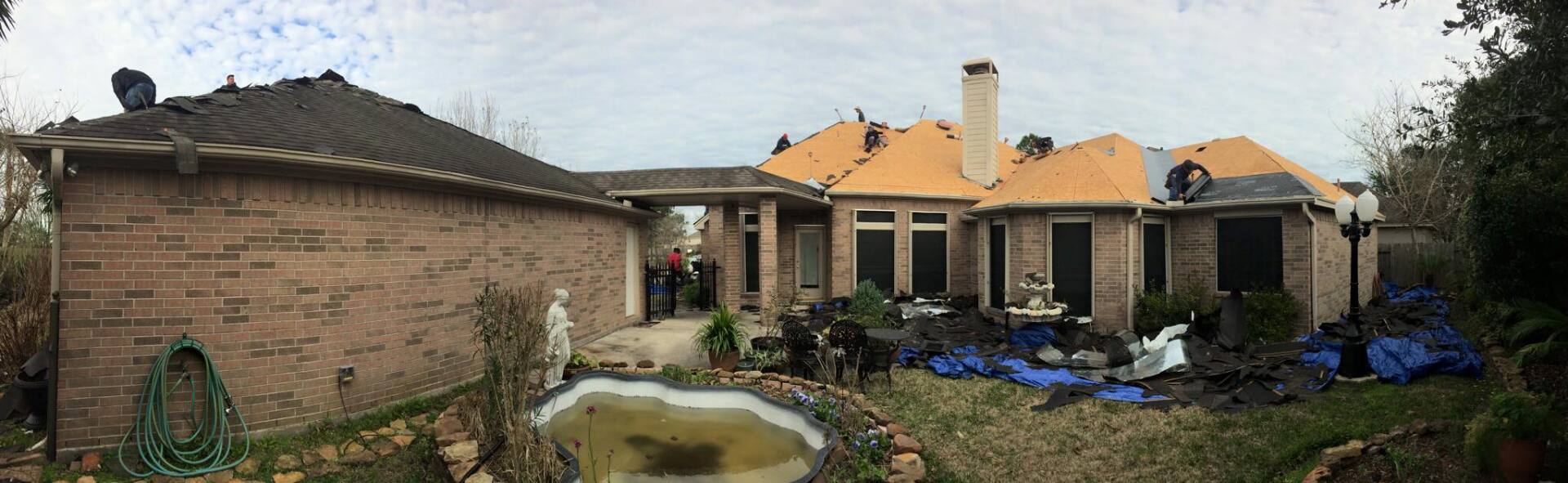 A brick house under renovation with workers on the roof, exposed wood decking, and a small backyard pond.