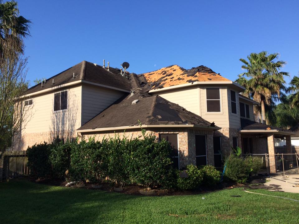 A two-story tan suburban house with significant roof damage and missing shingles, surrounded by shrubs and palm trees.