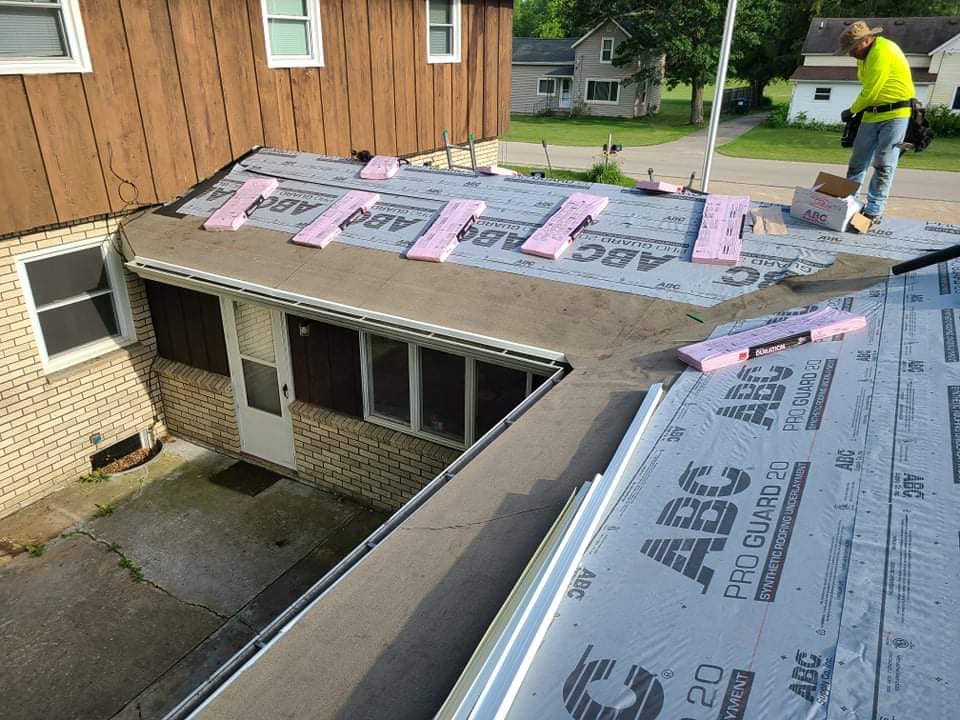 A construction worker in a high-visibility yellow vest installs insulation and roof underlayment on a flat house roof.