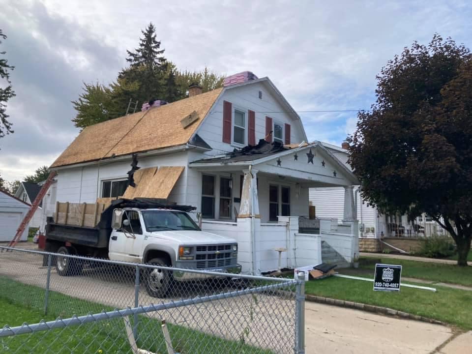 A white house under renovation with new plywood roof decking and a white dump truck parked in the driveway.