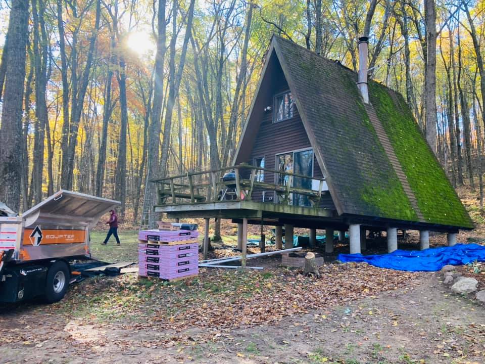 A brown A-frame cabin with a mossy roof in a wooded area, with a dump trailer and stacked purple pallets nearby.
