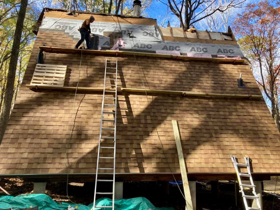 A person works on the roof of a house under construction in a wooded area, with scaffolding, ladders, and tarps visible.