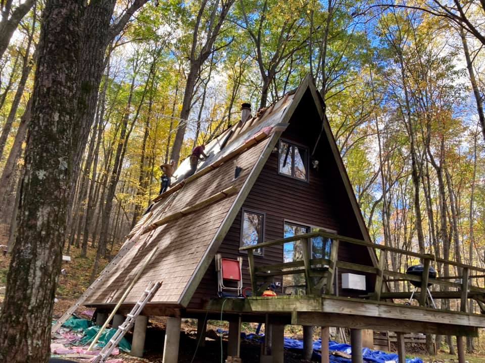 Two workers repair the roof of an A-frame wooden cabin set in a forest during autumn.