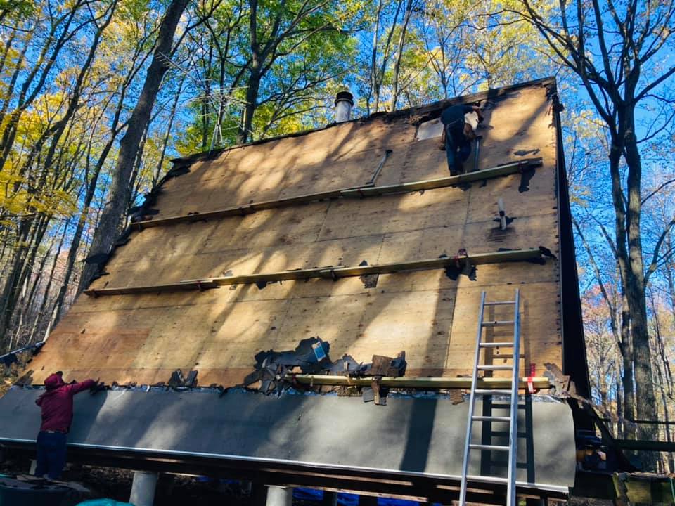Two workers on ladders install plywood panels on the steep roof of a cabin surrounded by trees.
