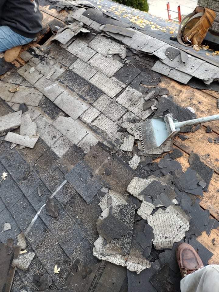 A worker using a metal tool to remove old asphalt shingles from a residential roof deck.