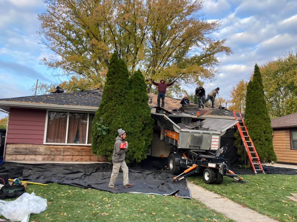 A crew works on a house roof with a lift parked by the front door, over a lawn covered in protective tarps.