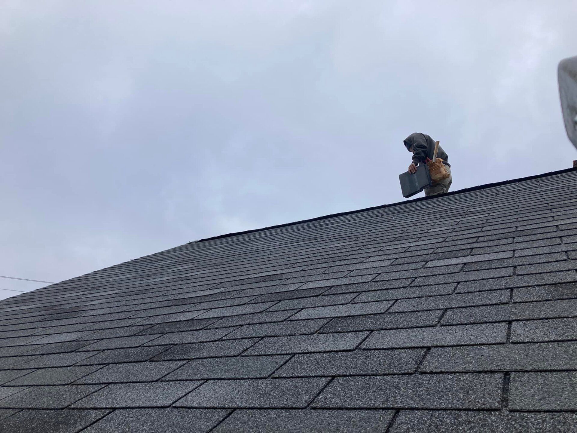 Roofer on a gray shingle roof working under a cloudy sky.
