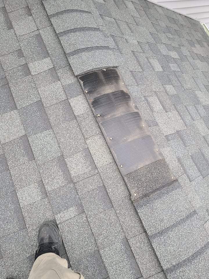 A section of gray asphalt shingle roofing with a missing row of ridge cap shingles, exposing the underlying vent material.