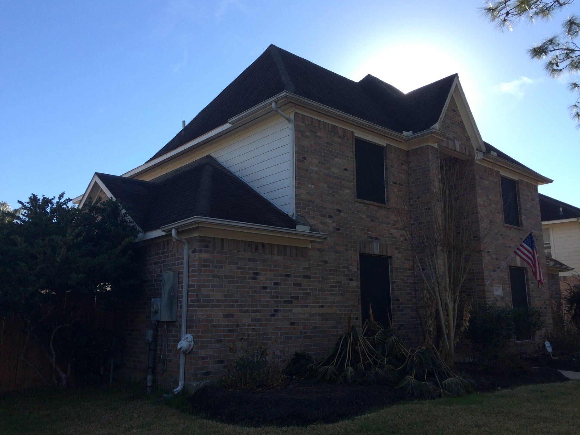 Two-story brick house with a dark roof and a small American flag hanging by the front entrance on a sunny day.