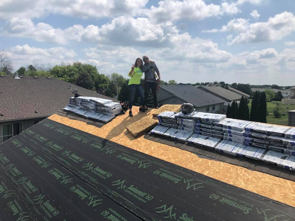Two people stand on a residential roof under construction, surrounded by stacks of shingles and underlayment.