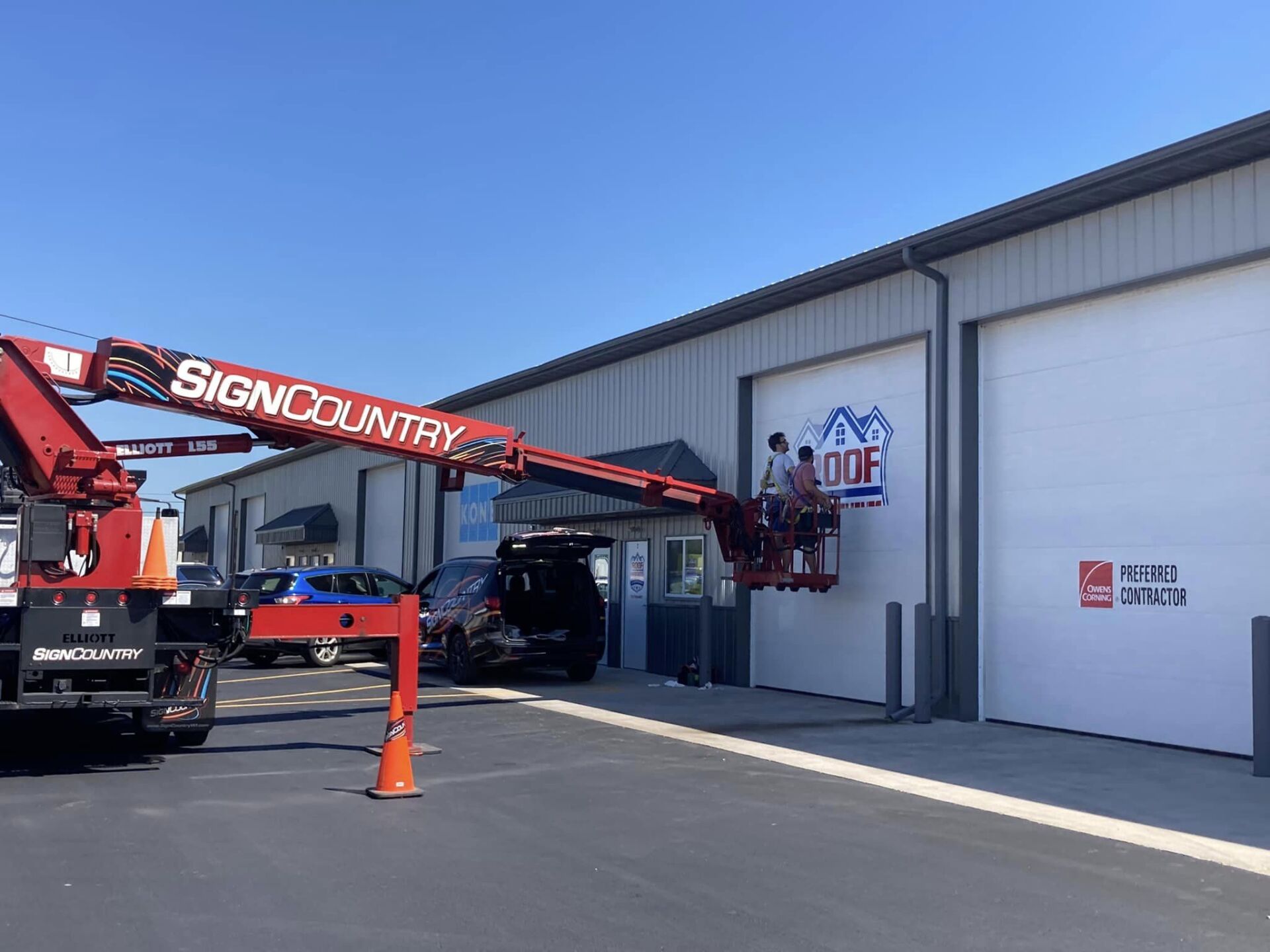 Workers in an elevated lift bucket apply a logo to the side of a gray metal building on a sunny day.