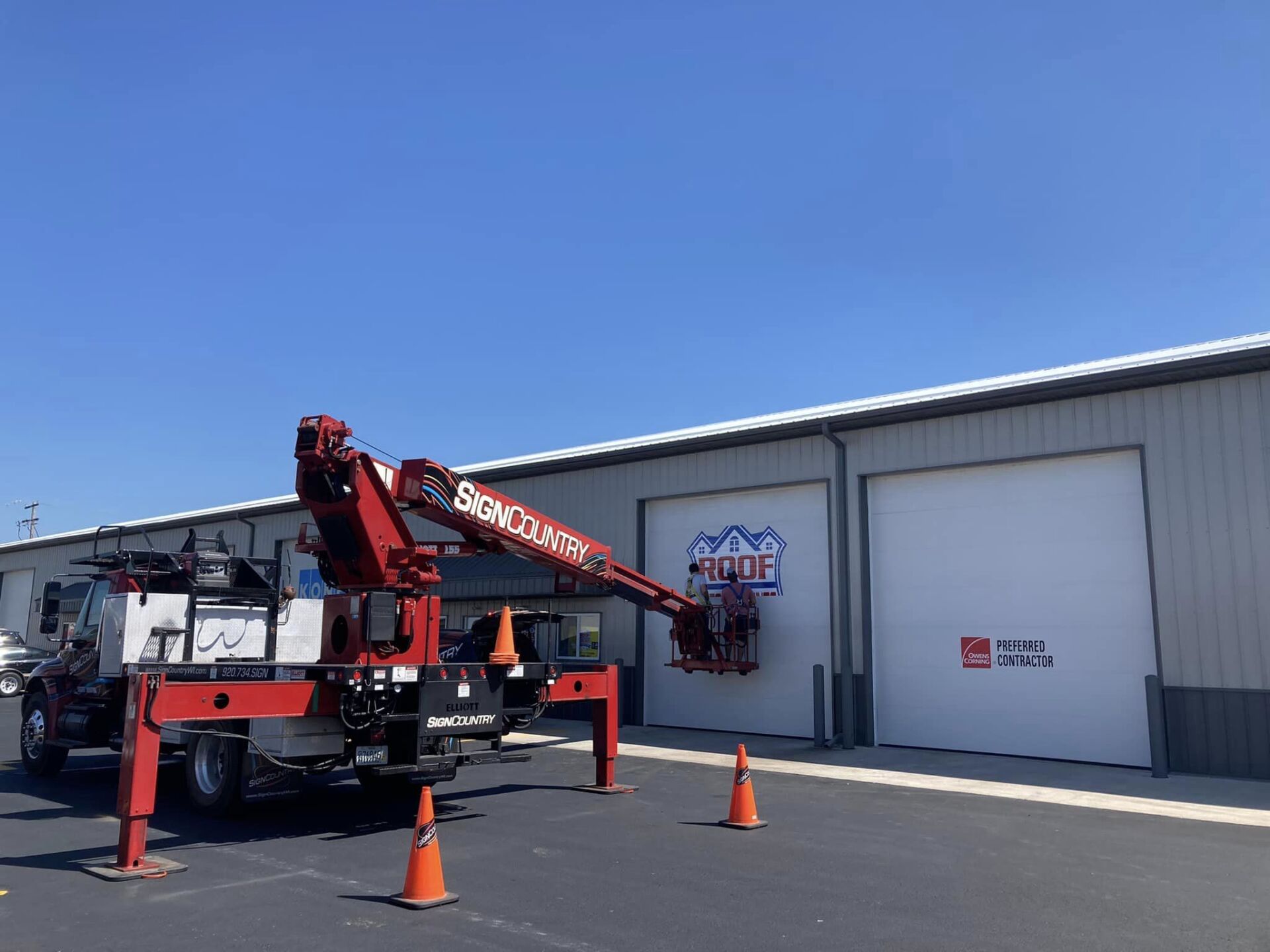 A red crane truck parked in a lot, with a worker in the bucket installing a sign on a warehouse door.