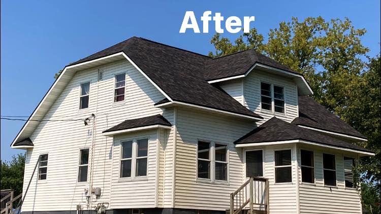 A two-story house with cream-colored siding and a newly installed dark shingled roof under a clear blue sky.