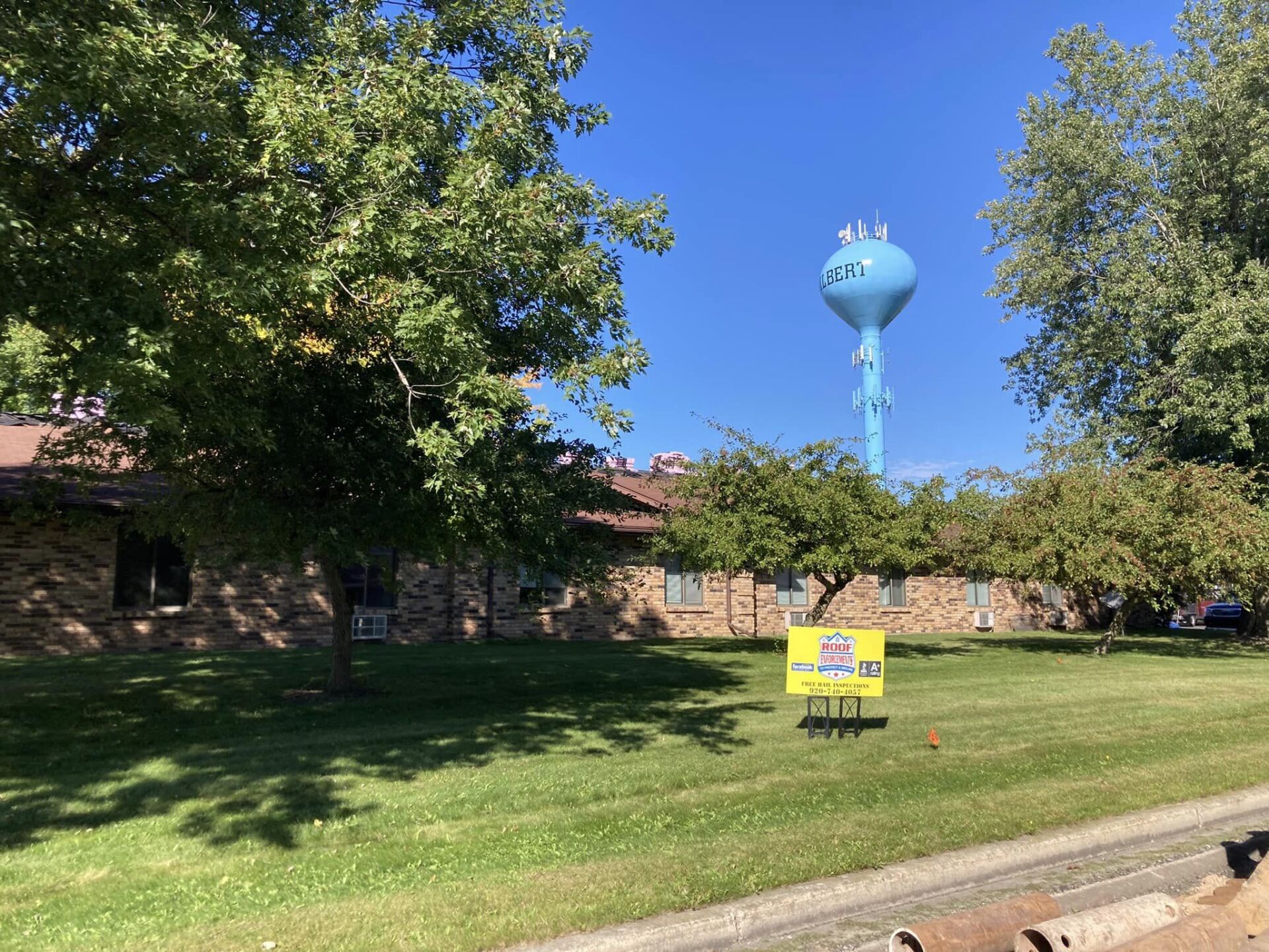 A brick building on a grassy lawn with a blue water tower under a clear sky and a yellow yard sign.