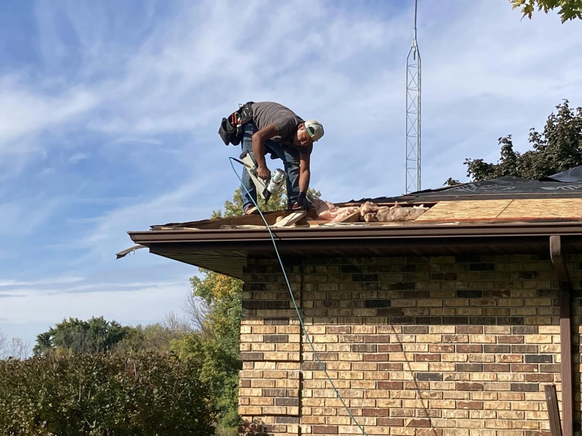 A construction worker uses a nail gun to install plywood sheathing on a roof against a blue sky.