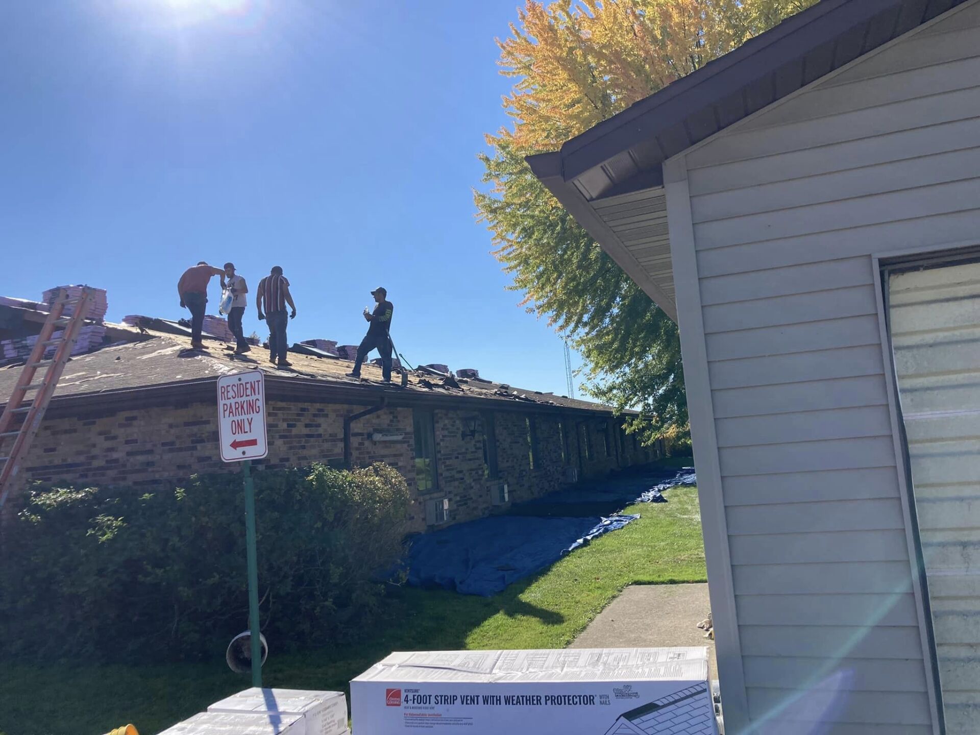 Four people work on a residential roof under a bright sunny sky, with a blue tarp on the lawn below.